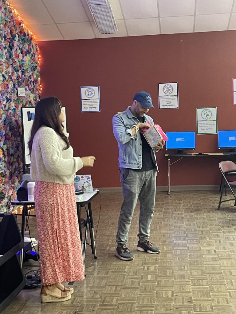 A man in a denim jacket and cap stands next to a woman in a sweater and long skirt, looking into a gift bag in a room with colorful decorations and computers, reminiscent of a student pitch event.