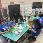 A group of people sits around a turquoise table, engaged in the VentureLab Idea to Pitch program while crafting vibrant paper creations. Posters and art adorn the wall behind them, adding inspiration to their innovative meeting.