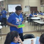 In the bustling Garcia Center, a person in a blue shirt holds a green item, possibly an idea to pitch, while standing in a room filled with tables, chairs, and people wearing matching shirts. A large mirror reflects the lively scene of this student pitch event.