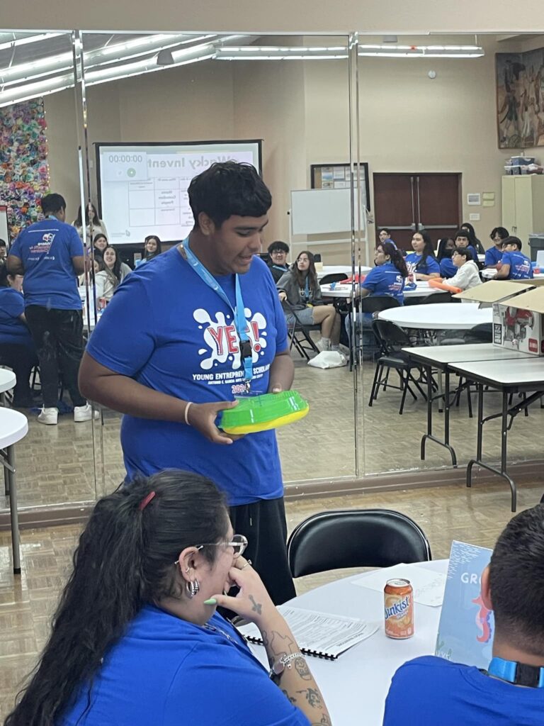 A young person stands holding a green container, proudly wearing a blue event t-shirt, in a room buzzing with ideas. This gathering is part of the VentureLab Idea to Pitch program implementation. A wall with mirrors and a screen reflects the anticipation as people are seated around tables.