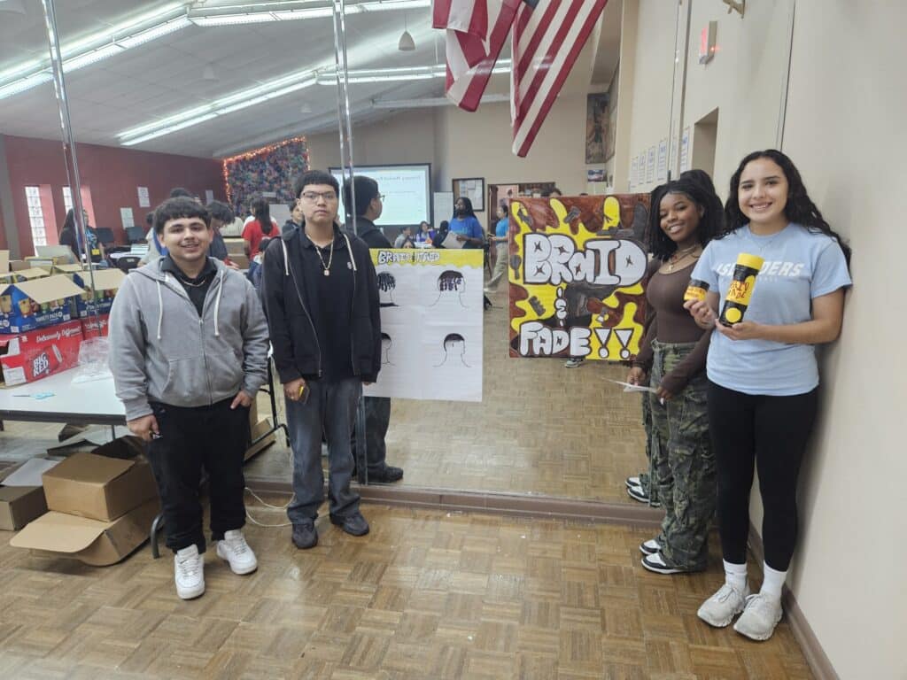 Four participants stand indoors holding hair-themed posters and a product, embodying the spirit of the VentureLab Idea to Pitch program. They are positioned by a wall adorned with mirrors and an American flag, while tables and boxes subtly frame this scene of innovation in the background.