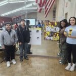 Four participants stand indoors holding hair-themed posters and a product, embodying the spirit of the VentureLab Idea to Pitch program. They are positioned by a wall adorned with mirrors and an American flag, while tables and boxes subtly frame this scene of innovation in the background.