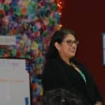 A woman stands in front of a colorful backdrop, smiling and wearing glasses and a black jacket. Behind her, a whiteboard and poster hint at the VentureLab Idea to Pitch program implementation, setting the stage for innovation.