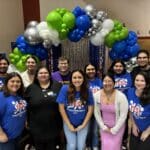 A group of people posing in front of a colorful balloon arch and shimmering curtain backdrop, some proudly wearing blue "YES!" shirts, captures the vibrant spirit of the Idea to Pitch student event.