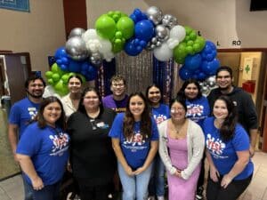 A group of people posing in front of a colorful balloon arch and shimmering curtain backdrop, some proudly wearing blue "YES!" shirts, captures the vibrant spirit of the Idea to Pitch student event.