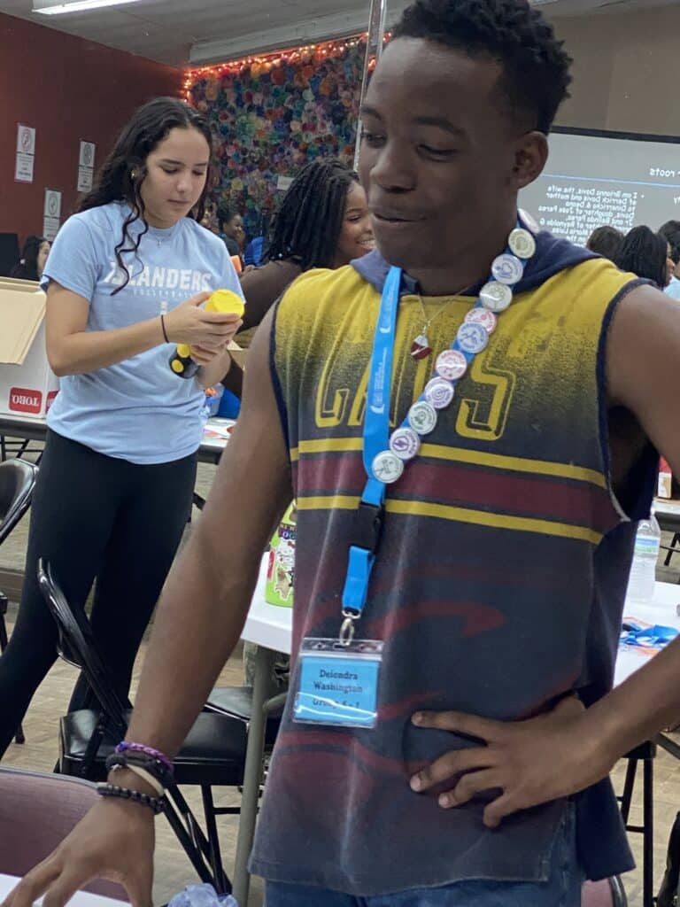 In a vibrant room, two people engage in animated discussions. One sports a colorful shirt and lanyard brimming with badges, while the other clutches a yellow item. The space, adorned with tables and decorations, buzzes with energy—ideal for exchanging ideas to pitch and fostering near-peer mentoring.