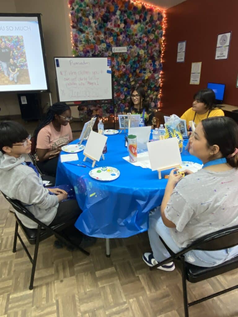 At the Garcia Center, a group gathers for the Idea to Pitch workshop. Around a table draped with a blue tablecloth, they paint on canvases, while behind them, a colorful wall and note-covered whiteboard fuel their creativity.