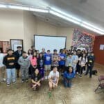 A diverse group of young people and adults gather at the Garcia Center, posing in a room with a projector screen and colorful wall. Some are seated while others stand, embodying the spirit of near-peer mentoring during the vibrant Spring Break Camp focused on youth entrepreneurship.