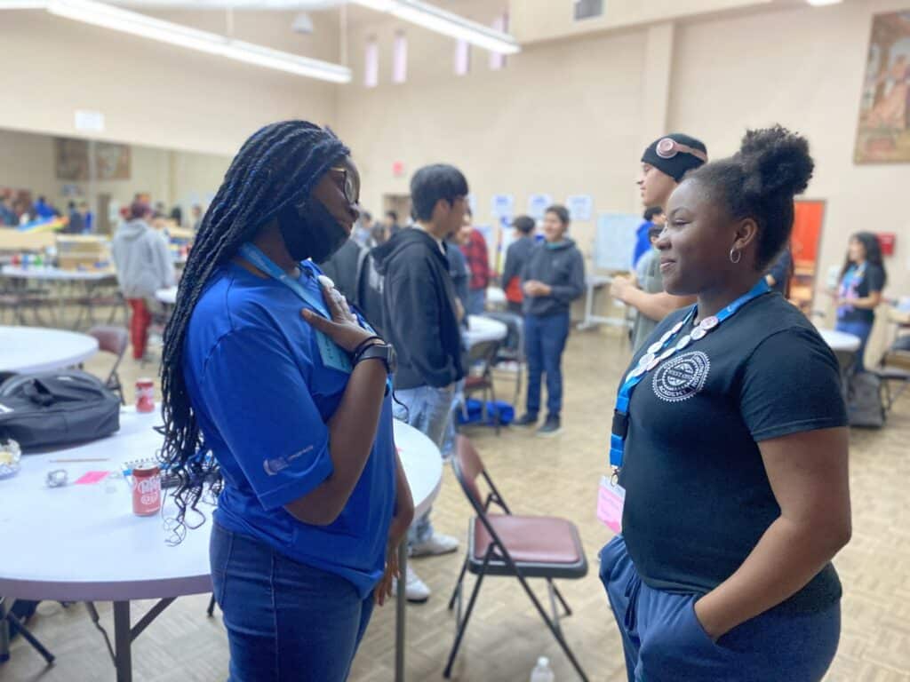 Two people stand and talk in a vibrant room bustling with activity. One wears a blue shirt and a mask, while the other sports a black shirt with a badge, discussing the exciting student pitch event at the Garcia Center Spring Break Camp for Youth Entrepreneurship.