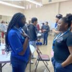 Two people stand and talk in a vibrant room bustling with activity. One wears a blue shirt and a mask, while the other sports a black shirt with a badge, discussing the exciting student pitch event at the Garcia Center Spring Break Camp for Youth Entrepreneurship.