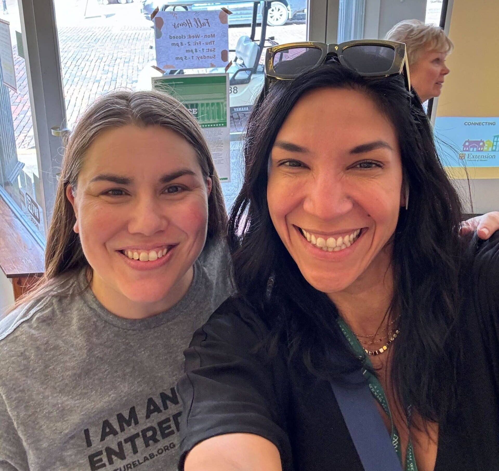 Two people smiling at the camera in an indoor setting, with one wearing a gray shirt and the other a black top, embodying the spirit of VentureLab collaboration. In the background, a person gazes out the window, symbolizing new horizons through education partnerships.