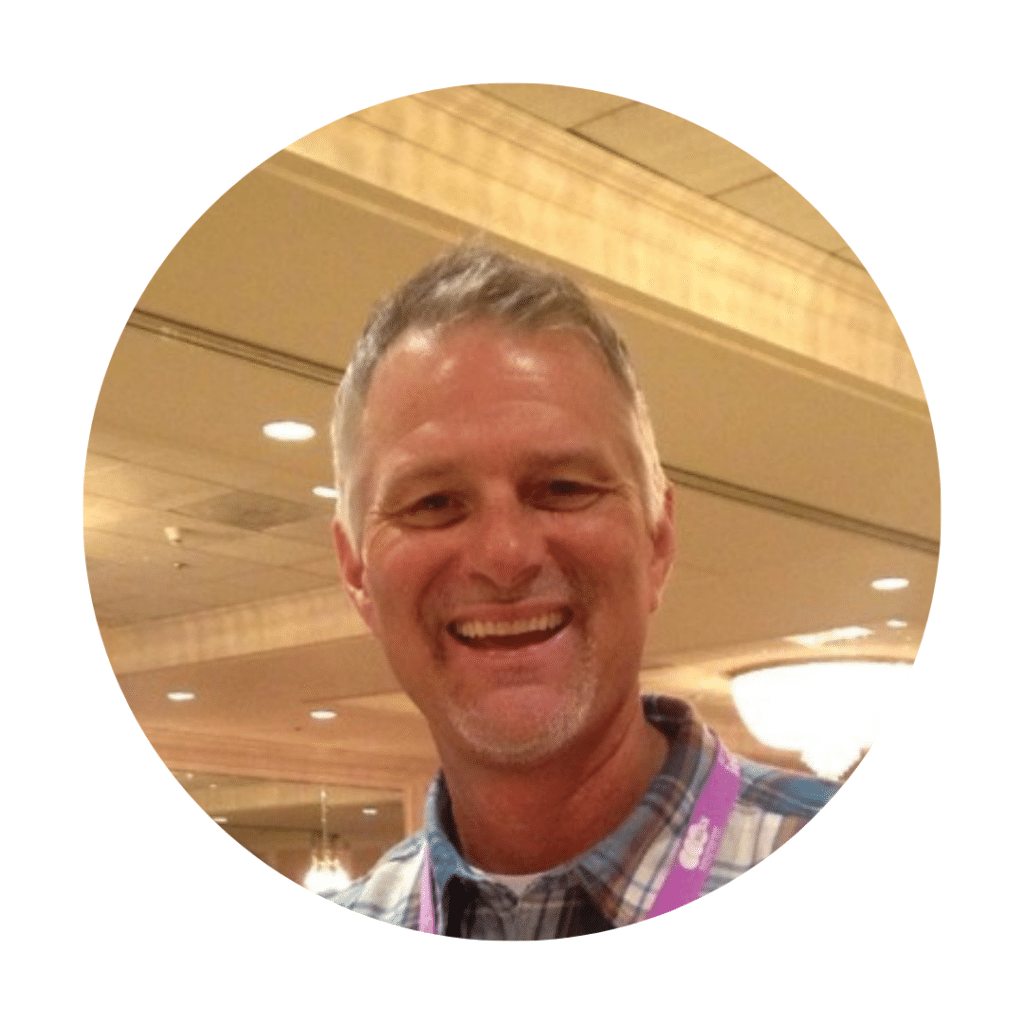A smiling man in a checkered shirt stands indoors, wearing a lanyard, eager to dive into the energy of VentureLab during STL Startup Week.
