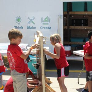 Children in red shirts engage in a hands-on activity outdoors, next to a mobile unit with "Think," "Make," "Create" signage. Sponsored by Nebraska’s Beyond School Bells initiative, this VentureLab inspires young minds to innovate and explore outside the traditional classroom setting.