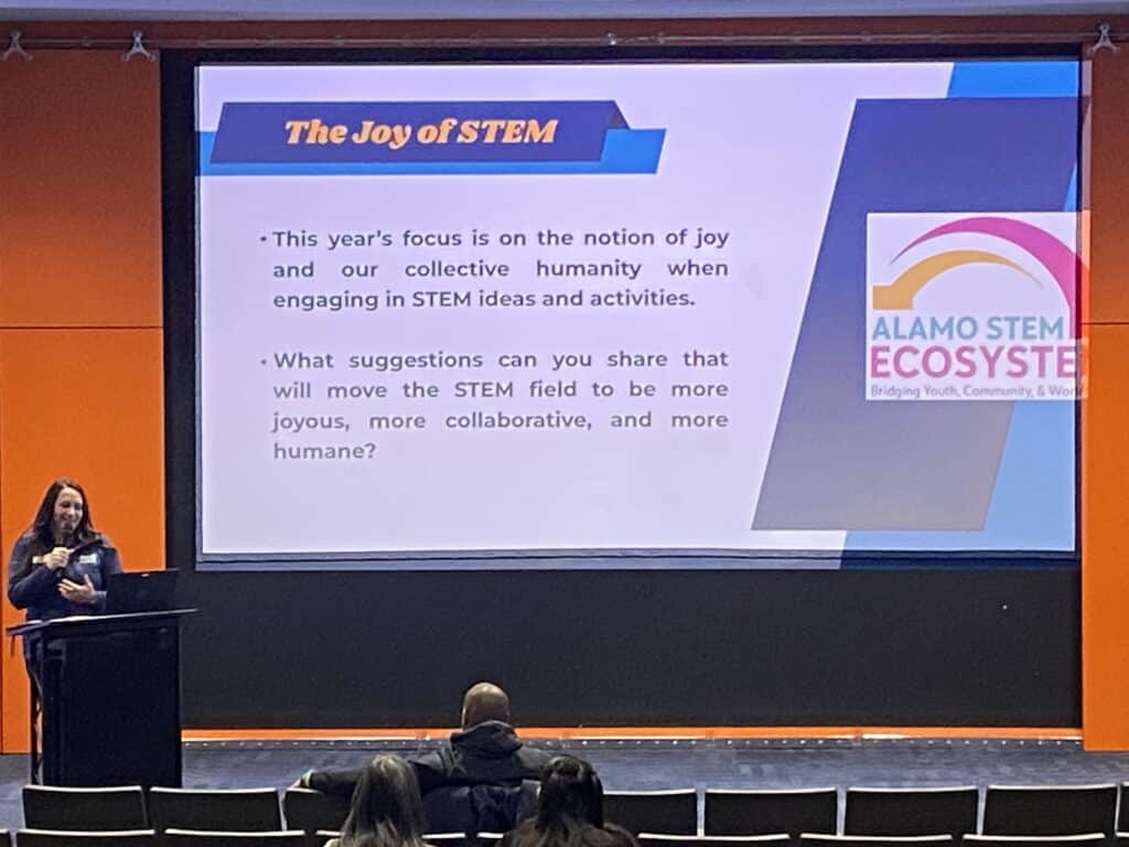 A presenter stands at a podium in front of a screen titled "The Joy of STEM," addressing an audience of San Antonio educators in an auditorium.